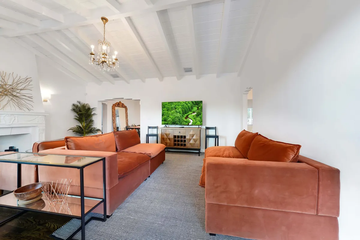 A cozy, modern lounge area with orange sofas, a prominently displayed modern television, a decorative mirror, lush green plant, and a chic chandelier. The space strikes a balance between vibrant style and comfort, perfect for unwinding or socializing.