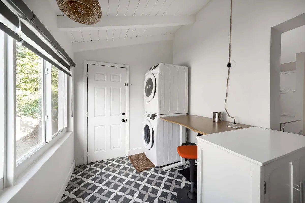 A cheerful laundry area with a full-size washer and dryer, a wooden table for folding clothes, and an orange stool adding a pop of color. Patterned floor tiles and natural light create a homey, practical space where daily chores feel easy and social.
