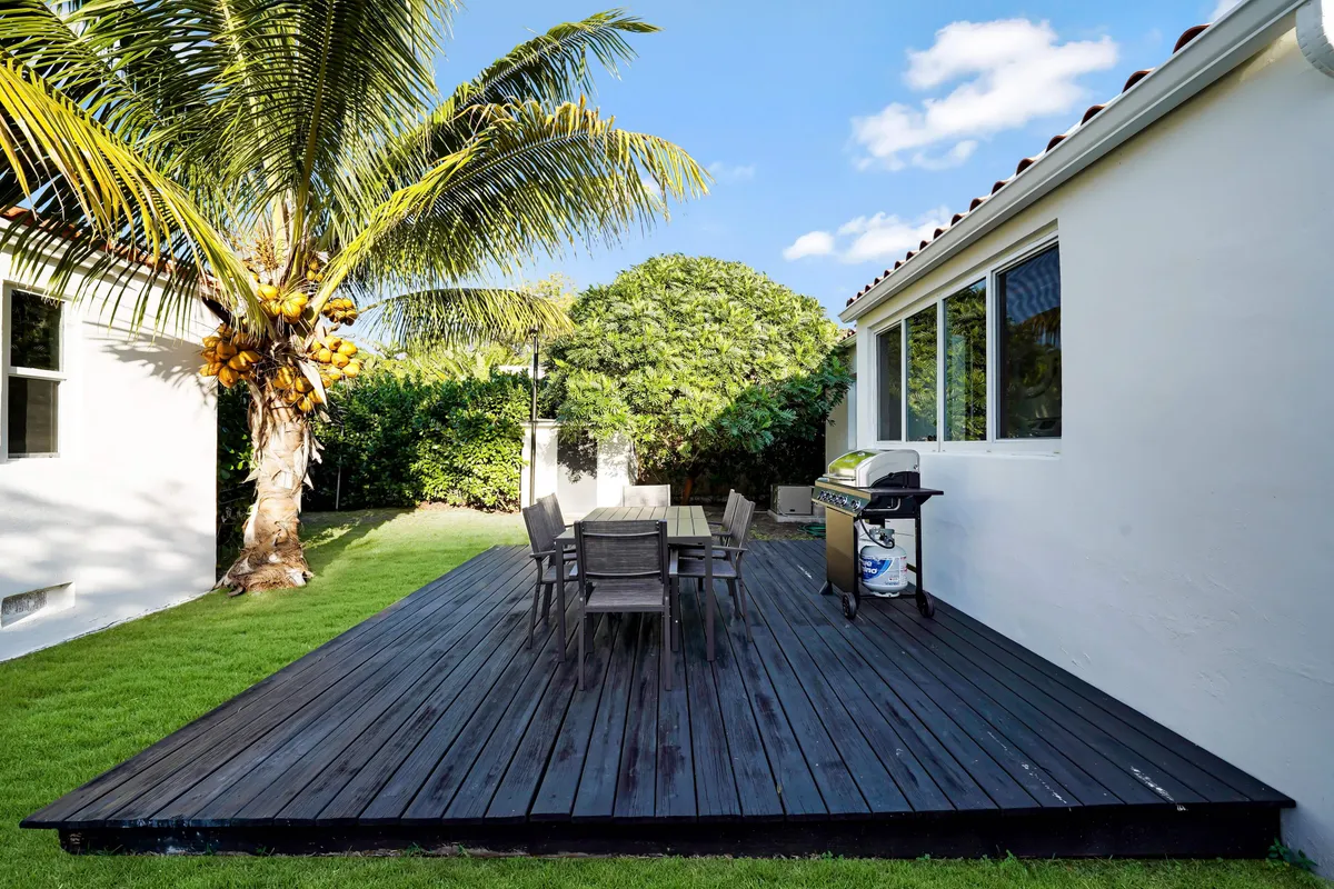 An inviting outdoor space featuring a wooden deck with a stylish dining table and grill, surrounded by lush palm trees. The setting is bright and open, ideal for relaxing in the sun, dining al fresco, or celebrating special occasions outdoors.