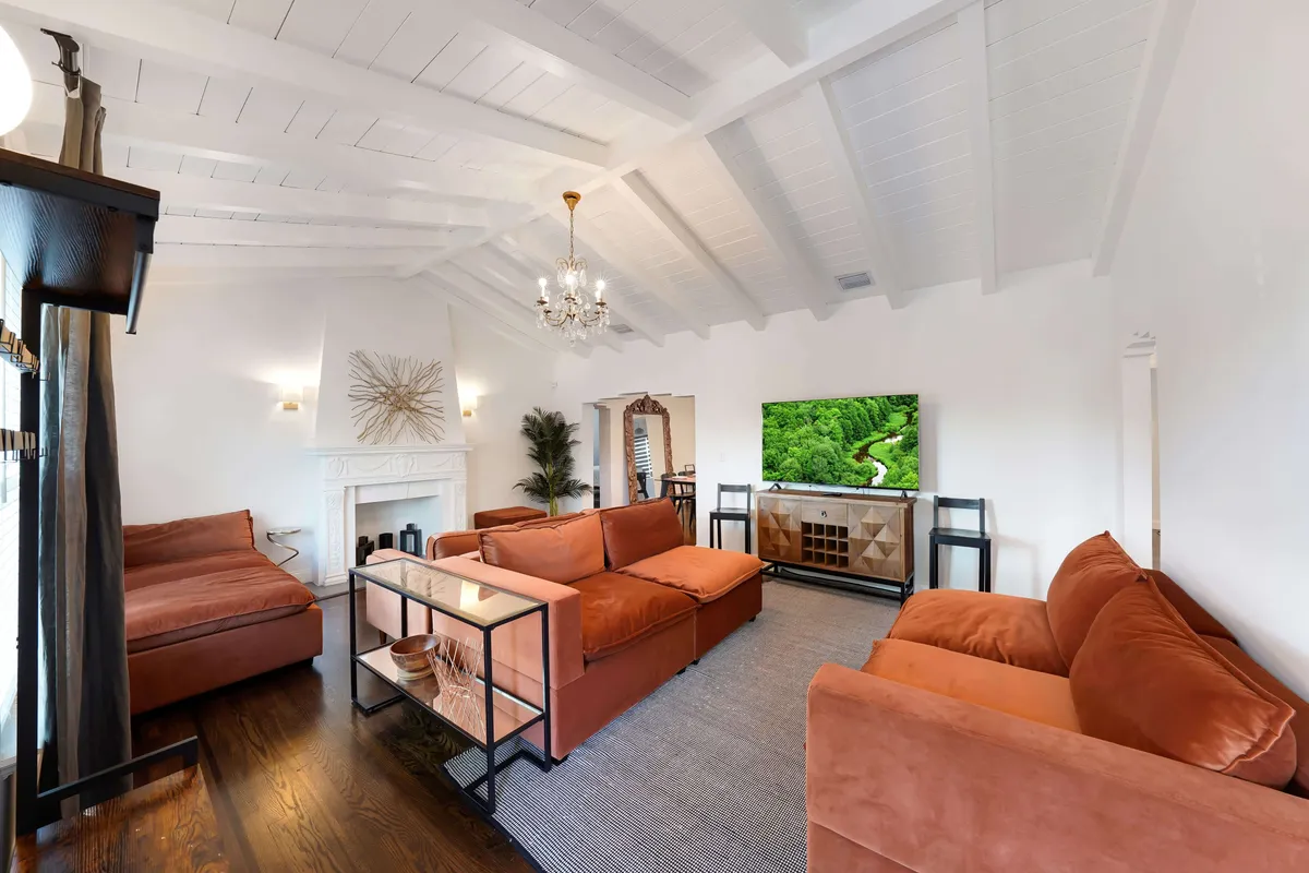 A cozy living room centered around a sofa, fireplace, television, and a statement chandelier. Multiple windows bring in natural light, creating a warm, inviting space for social gatherings or quiet relaxation.