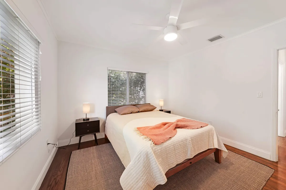 A serene bedroom with a large bed, nightstands, ceiling fan, and a generous window filling the space with natural light. The mood is calm and restful, emphasizing comfort and relaxation.