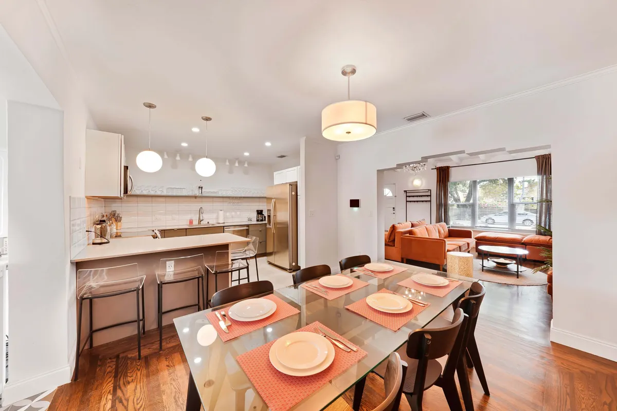 A spacious, open-plan dining and kitchen area featuring a table set for a meal, a kitchen island with bar stools, and a comfortable seating area with large windows that flood the space with natural light. The windows showcase the sun-filled aspect of the interior, while the room’s design feels both modern and welcoming.