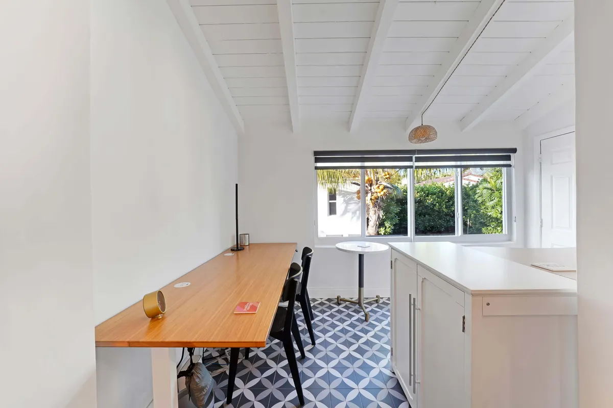 A bright, modern interior featuring a long wooden table and a round dining table set against a large window that fills the room with natural light. The space feels cozy yet open, suitable for both casual dining and work-from-home setups. There are clear indications of a comfortable, adaptable workspace within a serene environment.