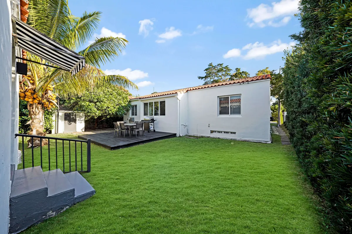 This image features a lush green lawn bordered by palm trees, with a dining table set up outside and the white exterior of the eco-retreat in view. The scene is tranquil and inviting, highlighting comfortable outdoor living surrounded by nature.