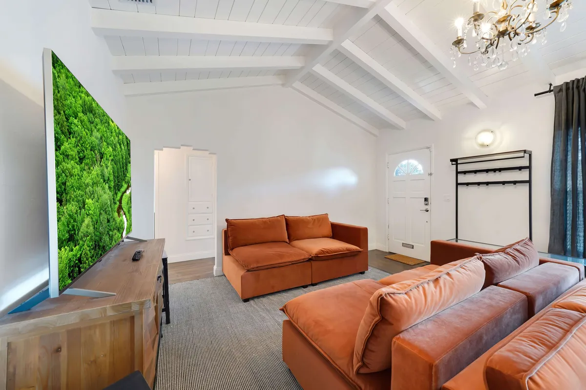 A welcoming, well-lit living room with a comfortable sofa, a prominent television, elegant chandelier, and a doorway that hints at the flow between living spaces. The space feels both relaxing and ideal for gatherings or quiet downtime.