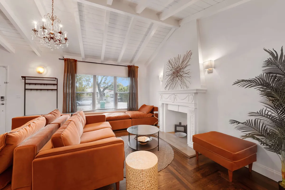 The image showcases a sunlit living room featuring a rust-colored sectional sofa, a modern white decorative fireplace topped with sculptural art, and a large window dressed with brown curtains. The space is accented by a round glass coffee table and designer furnishings, creating a cozy and stylish atmosphere ideal for relaxing or socializing.