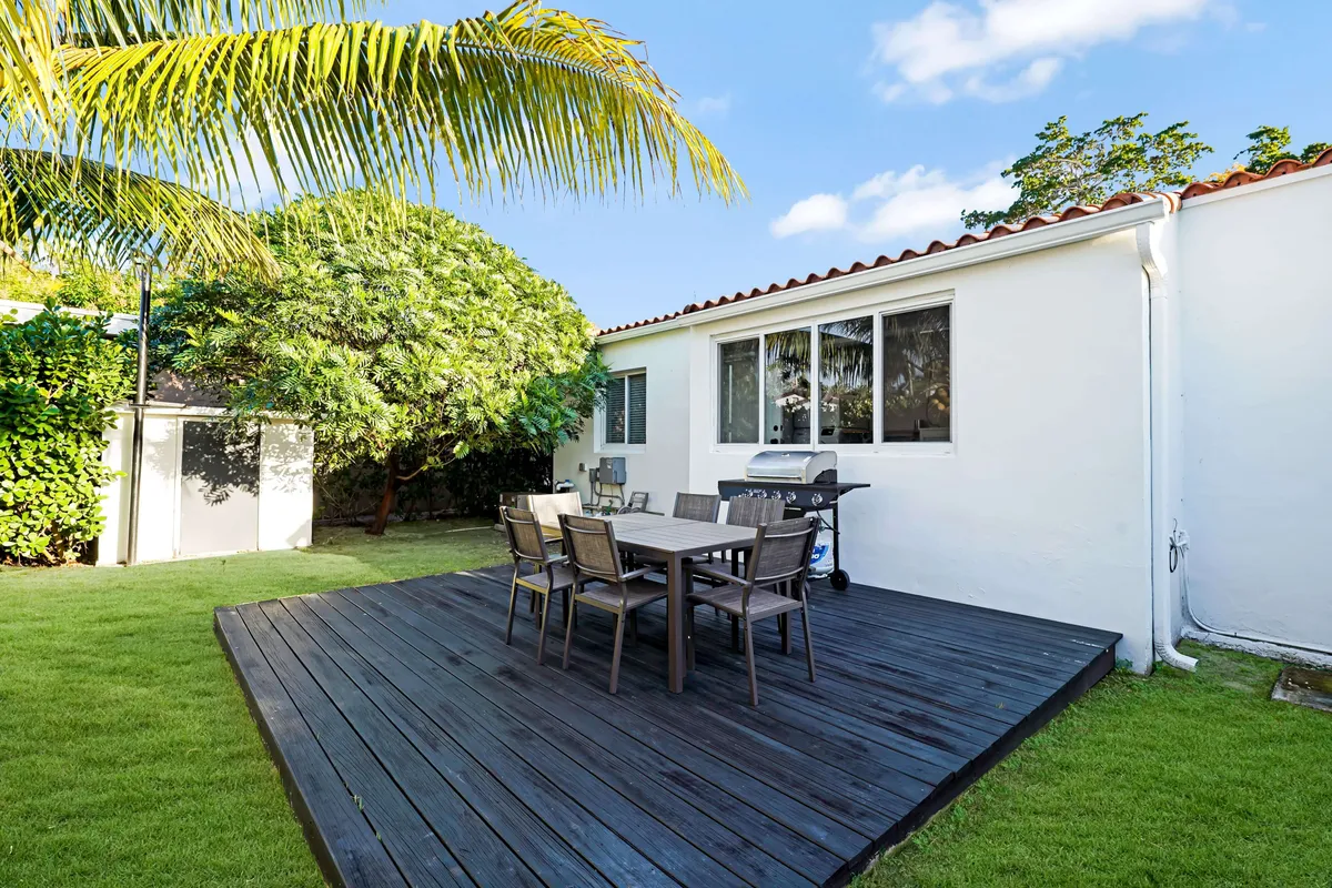 A spacious dark wooden patio deck offers an outdoor dining table with six chairs and a barbecue grill, set against a backdrop of lush tropical greenery and a vibrant green lawn. Palm trees sway nearby, creating a relaxed, social atmosphere perfect for enjoying Miami's outdoor lifestyle.
