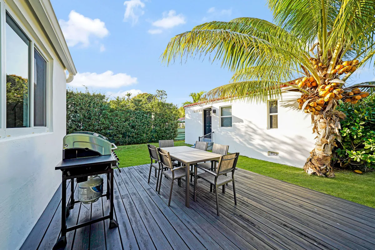 This image shows an inviting outdoor dining space featuring a table with chairs, a gas BBQ grill, a lush green lawn, and a tall palm tree. The atmosphere is leisurely, warm, and surrounded by greenery, making it ideal for relaxing or hosting gatherings in a private garden.