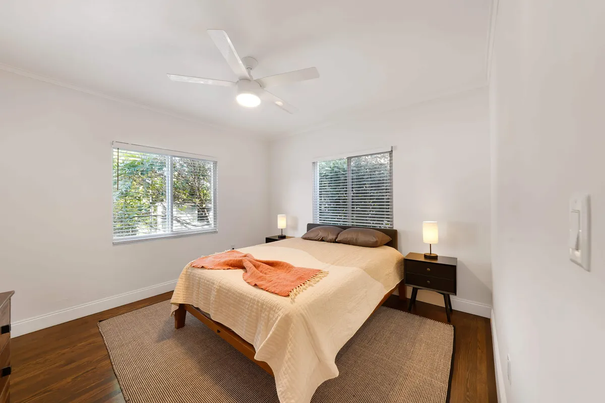 A peaceful bedroom with a beige-covered bed accented by an orange throw, two bedside tables with lamps, windows with white blinds, and a ceiling fan. The space is uncluttered, filled with gentle light, and invites relaxation.