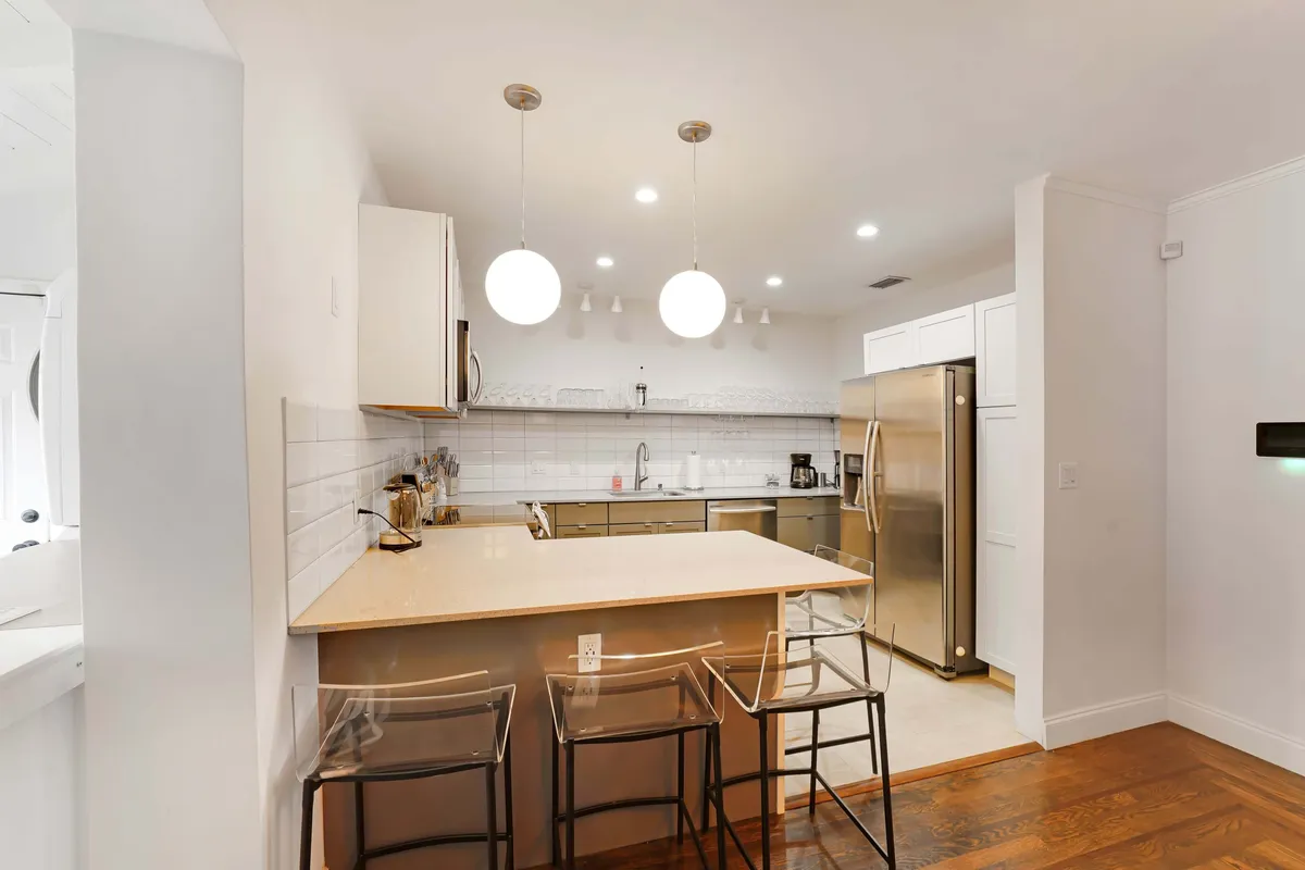 A sleek, modern kitchen with a central island featuring transparent bar stools, a stainless steel refrigerator, white cabinetry with a subway tile backsplash, and modern pendant lighting. The space is bright and efficient, with clear visibility of quality appliances and a dedicated coffee station area.