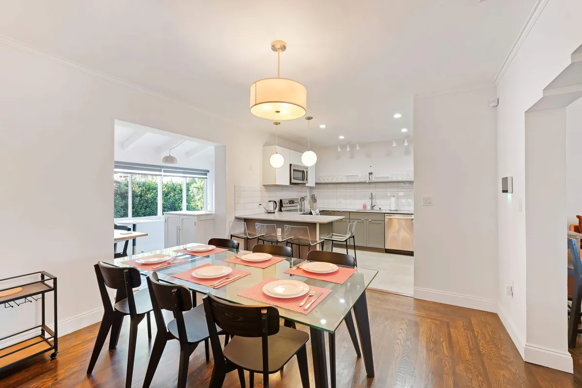 A bright, modern open-plan kitchen and dining area is shown, featuring a glass dining table elegantly set for six, a spacious kitchen island with bar stools, large sunlit windows, and contemporary appliances and cabinetry. The atmosphere is airy and welcoming, ideal for both family meals and casual gatherings.