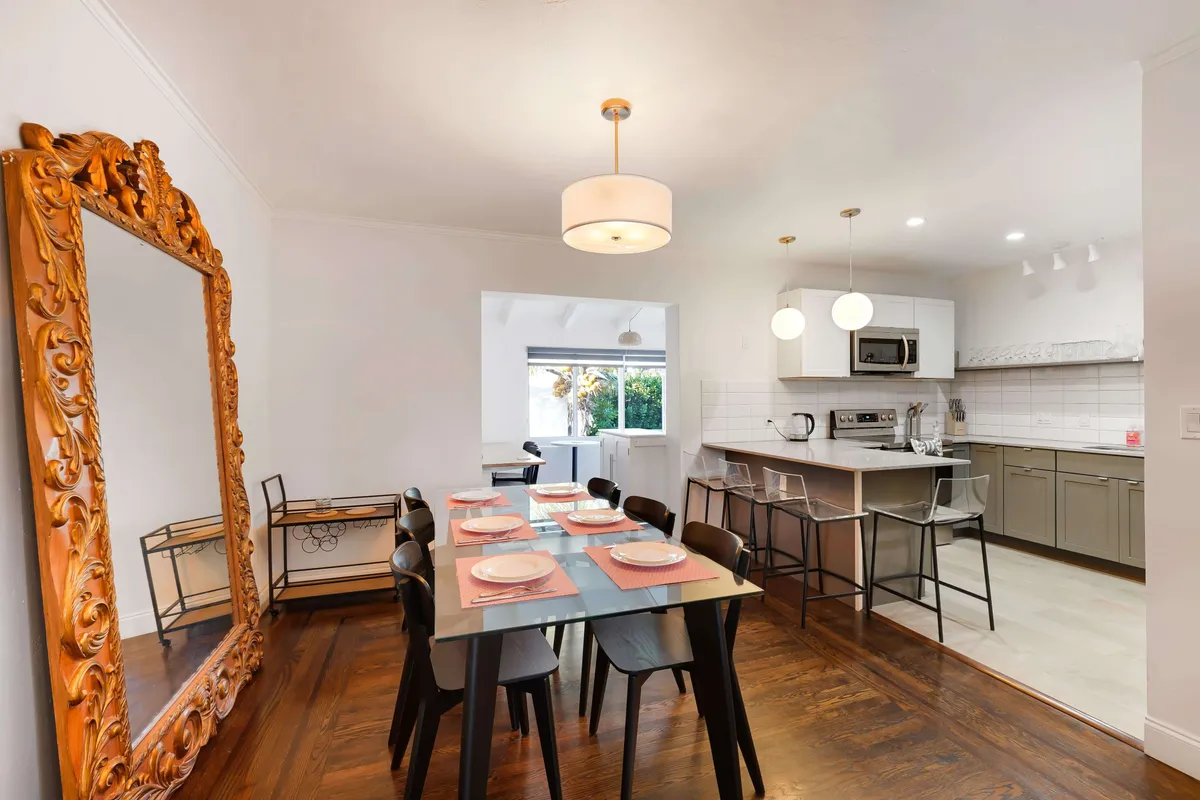 A warm, inviting view of an open kitchen and dining area, with a glimpse of the kitchen island and dining table set for six. The ornate mirror and pink placemats add a touch of comfort and style, suggesting a welcoming atmosphere ideal for breaks and creative thinking.