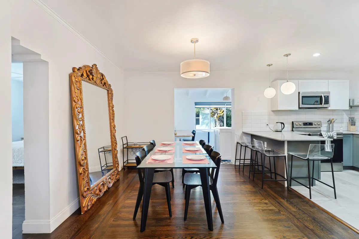 A modern, open-plan kitchen and dining area features a sleek black dining table set for eight, stylish pendant lighting, a kitchen island with bar stools, and stainless steel appliances. Bright white cabinetry and an ornate golden mirror create a welcoming, elegant space for family meals.