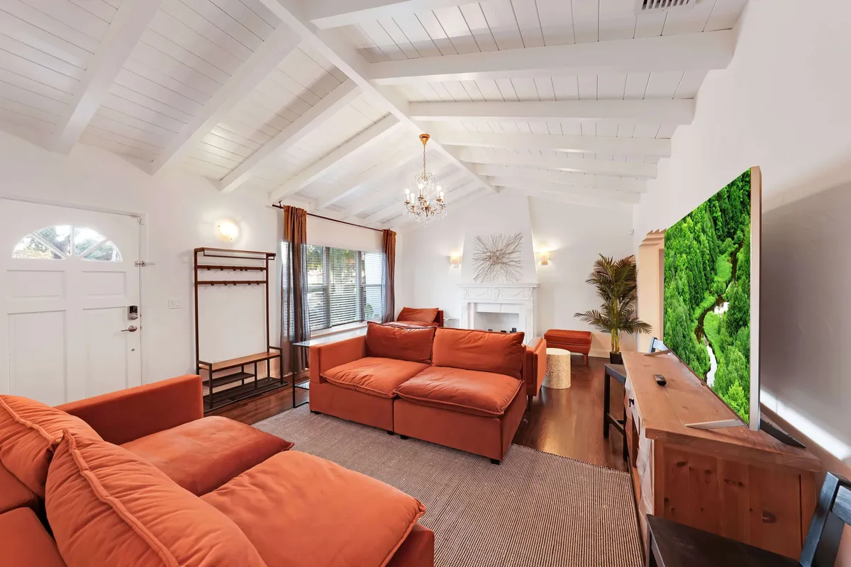 A cozy, modern living room with rust-colored sofas, an 8K Smart TV displaying a vivid nature scene, white vaulted ceiling with a chandelier, and a decorative fireplace. Large windows let in abundant natural light, highlighting the room’s inviting and sophisticated design.