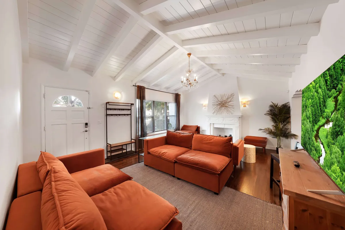 This image features burnt orange sectional sofas arranged for conversation, a white vaulted ceiling with exposed beams, a decorative fireplace with modern art above, and a large flat-screen TV. The space is flooded with natural light, giving it a warm, airy feel.