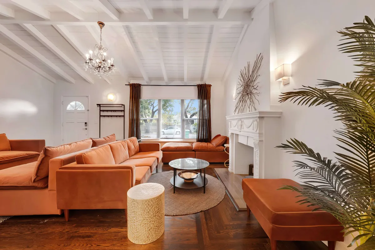 A spacious living room featuring a large burnt orange sectional sofa arranged around a glass coffee table and a round jute rug. An ornate white fireplace mantel anchors the space, while a chandelier hangs from the vaulted ceiling, accentuating the room's height. Natural light pours in, highlighting the blend of modern design with comfortable, inviting details.