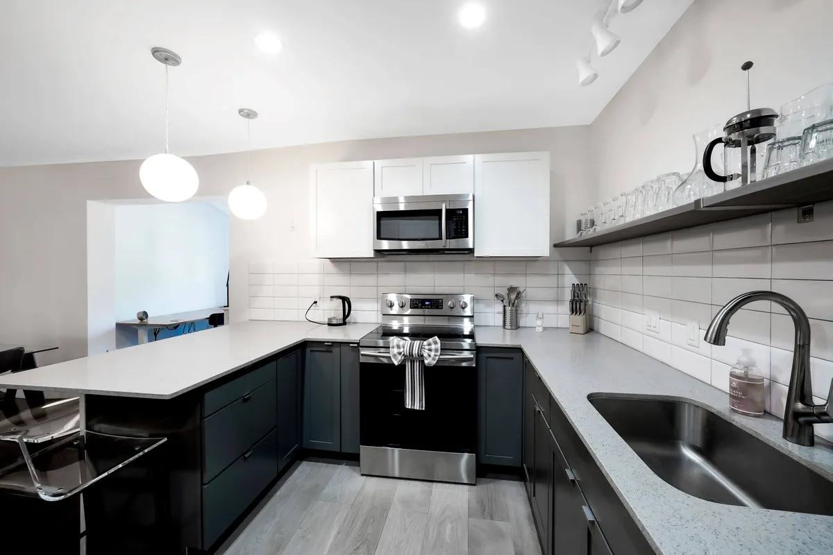 A well-organized kitchen showcases a stainless steel stove and microwave, large L-shaped countertop, modern sink, and open shelving with glassware and a coffee press. Natural light enhances the inviting and efficient layout, with clear signs of a fully stocked kitchen and coffee-making amenities.